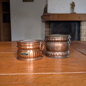 May include: Two antique copper containers with a hammered texture and brass accents sit on a wooden table. The containers have a cylindrical shape with handles and decorative details. One has a small plaque.