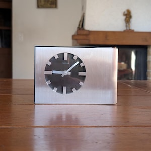 May include: A brushed metal desk clock with a black clock face and silver hands. The clock has a rectangular base and a circular clock face with white markers. The clock is sitting on a wooden table.