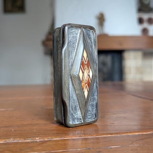 Op de afbeelding: Een rechthoekige, zilverkleurige sigarettenetui met een ruitvormige inleg van rode, oranje en crèmekleurige stenen. De etui heeft een getextureerd, geëtst ontwerp en staat rechtop op een houten tafel.