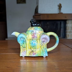 Op de afbeelding: Een kleurrijke keramische theepot met een zwart kattenbeeldje op het deksel. De theepot heeft een gele basis met bloemen- en krulontwerpen in blauw, groen en roze. De handgreep en tuit zijn geel. De theepot staat op een houten tafel.