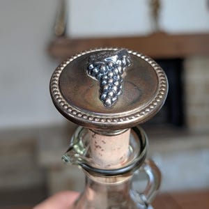 Pode incluir: Uma rolha de garrafa de vidro transparente com um topo metálico circular com borda de contas e um design em relevo de cacho de uvas prateado no centro. A rolha inclui uma base de cortiça natural que se encaixa no gargalo da garrafa.
