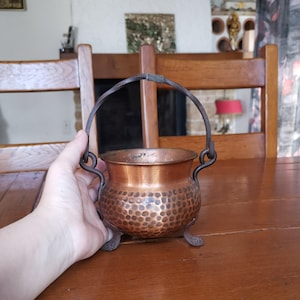 Puede incluir: Un pequeño caldero de cobre martillado con un asa de hierro forjado y tres patas decorativas. La olla de cobre tiene un color rico y cálido y una superficie texturizada. El asa está unida a la olla con bucles decorativos.