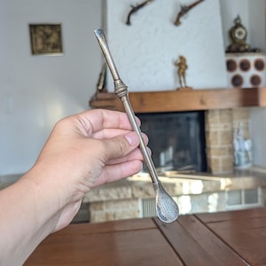 May include: A silver-toned metal bombilla straw featuring a slender, tubular stem with a decorative ribbed ring near the top and a flattened, perforated spoon-shaped filter at the bottom end, designed for filtering loose leaf tea infusions.