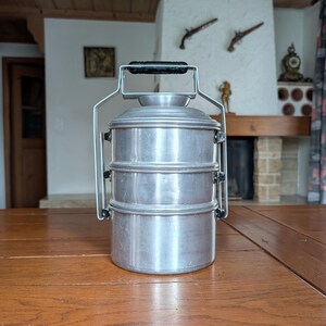 May include: A three-tiered, silver metal lunch carrier with a black handle. The carrier has a handle for easy transport. The lunch carrier is on a wooden table. The background includes a fireplace and wooden ceiling.