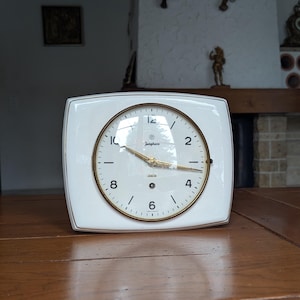 May include: A white, square-faced Junghans clock with a gold-tone circular clock face. The clock has gold hands and a gold rim. The clock sits on a wooden table. The background includes a fireplace and decorative items.