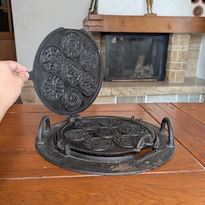 May include: A black cast iron krumkake or waffle iron featuring a circular design with seven distinct embossed floral and geometric patterns on both the top and bottom plates, equipped with two side handles and a hinged mechanism for opening.