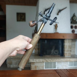 May include: An antique brass and metal blowtorch with a ribbed handle and various nozzles and valves. The torch is held against a background of a fireplace and wooden table.
