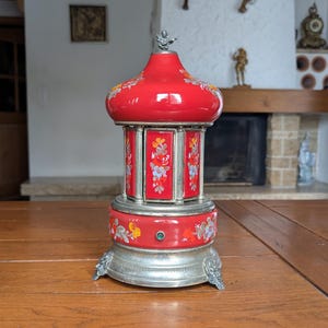 May include: A red carousel-style music box featuring hand-painted floral motifs in orange, white, and blue. The structure includes a metallic silver base with ornate feet, a central rotating section with vertical panels, and a domed top with a small finial.