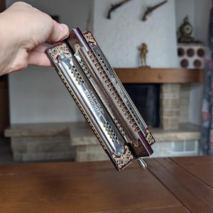 May include: Two vintage harmonicas with silver and gold accents. The harmonicas have a dark brown wooden frame with ornate gold designs. One harmonica reads "TREMOLO harmonica". The other harmonica reads "M. Hohner".