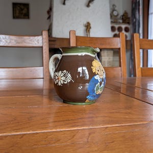 Pode incluir: Um jarro de cerâmica castanho com uma pega branca e uma borda verde. O jarro apresenta um desenho floral e uma ilustração pintada de uma pessoa com roupa azul. O jarro está numa mesa de madeira.