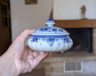 May include: A white and blue ceramic trinket box with a lid. The box features a floral design in blue and is held in a hand. The box is approximately 10 cm in diameter and 8 cm tall. The lid has a small finial.