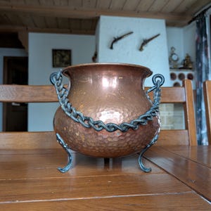 May include: A hammered copper pot with a decorative chain handle and three curved feet. The pot has a rich, warm copper color and a textured surface. The chain and feet are a dark, contrasting metal.
