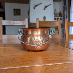 May include: A small, hammered copper pot with a dark metal handle. The pot sits on a wooden table. The pot has a rounded shape and a textured surface. The background includes wooden chairs and a fireplace.