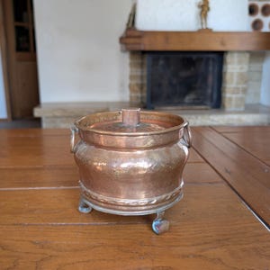 May include: A copper-colored, antique pot with a lid and small handles. The pot sits on three small feet. The pot is on a wooden table. The background includes a fireplace and a framed picture.