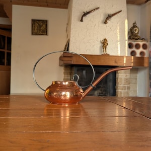 Puede incluir: Una regadera de cobre con un pico largo y un asa curvada. La regadera tiene un cuerpo redondeado y está sobre una mesa de madera. El fondo incluye una chimenea y elementos decorativos.