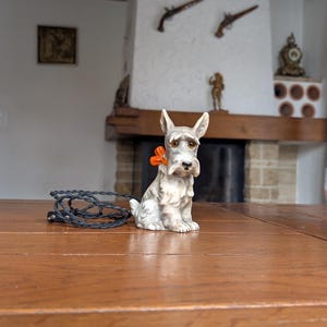 Könnte beinhalten: Eine Keramikfigur eines Scottish Terriers mit grau-weißem Fell, die eine orangefarbene Schleife trägt. Neben dem Hund ist ein schwarzes Kabel aufgerollt. Die Figur steht auf einem Holztisch.