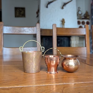 May include: Three copper containers on a wooden table. The largest is a bucket with a handle, the middle one is a small pot, and the smallest is a round bowl. The copper has a hammered texture. The table is made of wood.