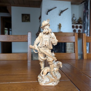 May include: A detailed, beige-colored figurine of a hunter with a rifle, binoculars, and a dog. The hunter wears a hat and period clothing. The dog stands at the hunter's feet on a decorative base. The figurine is set on a wooden table.
