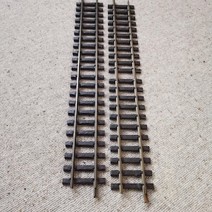 Puede incluir: Dos vías de tren modelo paralelas con traviesas de madera marrón oscuro y raíles de color latón. Las vías están colocadas sobre una superficie clara y texturizada, lo que sugiere un tema de modelismo ferroviario.