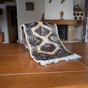 May include: A small, rectangular rug with a cream-colored background and a repeating pattern in black, red, and pink. The rug has a decorative fringe along the bottom edge. The rug is placed on a wooden table.