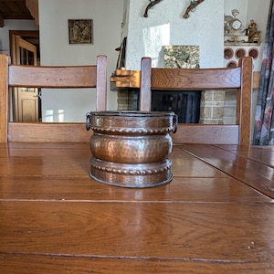 Puede incluir: Una jardinera antigua de color cobre, con textura martillada y dos asas pequeñas. La jardinera tiene forma bulbosa con un diseño de cuentas en la base. Se encuentra sobre una mesa de madera en una habitación con chimenea.