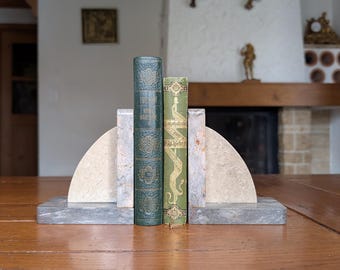 Puede incluir: Un juego de sujetalibros de mármol con base gris y lados semicirculares, que sostiene dos libros antiguos con cubiertas verdes y azules. Los libros están sobre una mesa de madera.