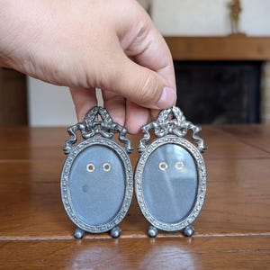 May include: Two antique silver-toned oval picture frames. Each frame has a decorative top with a bow and laurel leaves. The frames have a detailed border and small feet. The frames are being held up by a hand.