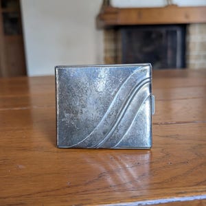 May include: A square, silver-toned metal cigarette case with a textured surface and a curved, wave-like design. The case is closed with a small clasp on the side. The case is sitting on a wooden table.