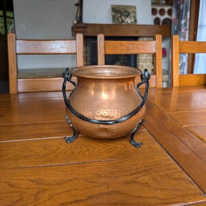 May include: A copper cauldron with a hammered finish, complete with black metal handles and decorative feet. The pot is displayed on a wooden table, highlighting its warm, metallic hues. This vintage-style pot is suitable for decorative use.