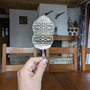 Puede incluir: Utensilio de servir plateado ornamentado con un diseño calado decorativo. El utensilio tiene un mango y una forma simétrica de dos niveles. El fondo incluye una mesa y sillas de madera, y una chimenea.
