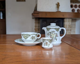 May include: A vintage tea set with a white background and a floral pattern in green, yellow, and pink. The set includes a teapot, a teacup with a saucer, and a creamer. The set is placed on a wooden table.