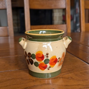 May include: A ceramic pot with a beige base, green rim, and two small handles. The pot is decorated with orange and yellow floral designs and brown leaves. The pot is sitting on a wooden table.