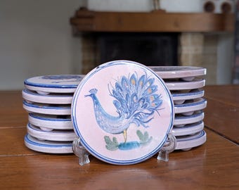 May include: A stack of round ceramic coasters with a peacock design. Each coaster features a light pink center with a blue peacock illustration and a blue border. The coasters are stacked on a wooden table.