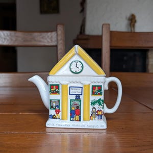 Può includere: Una teiera in ceramica bianca a forma di stazione ferroviaria, con tetto giallo e orologio. La teiera presenta illustrazioni di persone e il testo "Primrose Junction" e "Cafe". La base della teiera recita "a railway station teapot".