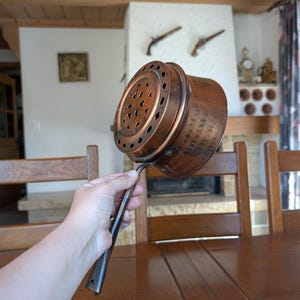 May include: An antique copper cooking pot with a long handle. The pot has a perforated top and a rounded body. The handle is dark and appears to be made of iron. The pot is held up in front of a wooden table and chairs.