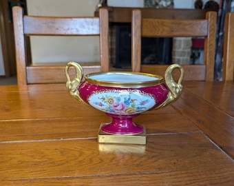 May include: A decorative, antique porcelain bowl with a burgundy base and gold accents. The bowl features a floral design and two gold handles. The base is square and gold. The interior rim is gold.