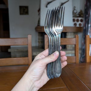 May include: A collection of antique silver forks, held in a hand. The forks have a tarnished appearance, with detailed patterns on the handles. The tines are long and slender, suggesting a vintage dining set.