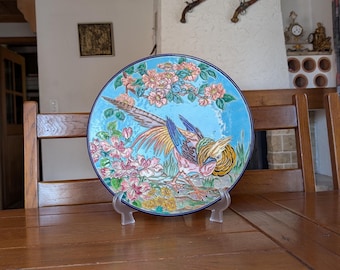 Pode incluir: Um prato decorativo de cerâmica com um faisão colorido rodeado por flores e folhagem rosa sobre um fundo azul claro. O prato é exibido em um suporte transparente. É um item decorativo.