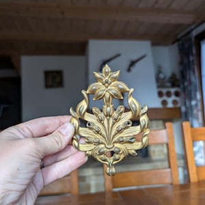 Puede incluir: Pieza decorativa de metal dorado con diseño floral y de hojas. La pieza simétrica tiene una flor central y elementos curvos y fluidos. Parece ser un artículo antiguo o vintage, posiblemente un adorno de mueble o elemento decorativo.