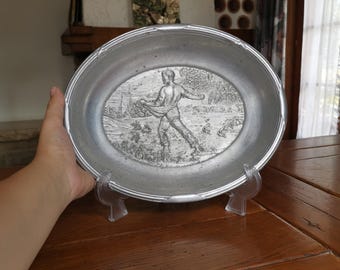 May include: Oval pewter plate with a detailed relief scene of a figure in a landscape. The plate has a decorative rim and is displayed on a clear plastic stand. The plate is likely vintage or antique.