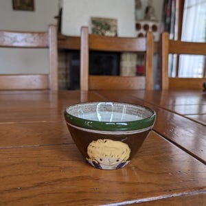 Pode incluir: Uma pequena tigela de cerâmica marrom com uma borda verde e uma ilustração de desenho animado de uma pessoa. A tigela está em uma mesa de madeira. O interior da tigela é de cor clara.