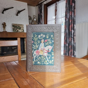 May include: A framed needlepoint artwork featuring a person on a white horse, surrounded by floral patterns. The artwork is set within a decorative, ornate silver frame. The background of the needlepoint is a teal color.