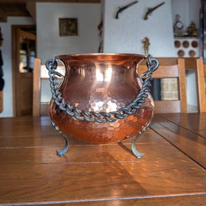 May include: A copper cauldron with a hammered finish, featuring a chain handle and three curved feet. The pot reflects light and sits on a wooden table. This vintage piece is likely for cooking or decorative purposes. The cauldron's dimensions are approximately 25 cm in diameter.