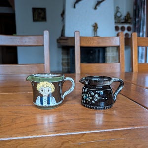 Pode incluir: Duas pequenas jarras de cerâmica sobre uma mesa de madeira. Uma jarra é marrom com uma ilustração pintada de uma pessoa. A outra é preta com um desenho floral. Ambas têm alças e bicos.
