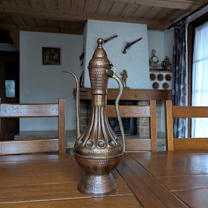 May include: A copper-colored decorative pitcher with a long spout and a handle shaped like a snake. The pitcher has a textured surface and a bulbous body. The pitcher is sitting on a wooden table.