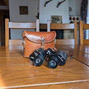 May include: Black binoculars and a brown leather case on a wooden table. The binoculars are marked "Kero" and "Armee Modell". The case is fastened with a buckle. The background features wooden chairs and a fireplace.