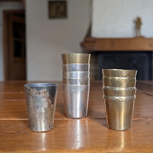 Puede incluir: Una colección de vasos de chupito de metal vintage en varios acabados. Los vasos incluyen plata deslustrada, plata pulida y latón. Los vasos de chupito están dispuestos sobre una mesa de madera, mostrando sus diferentes texturas y colores.