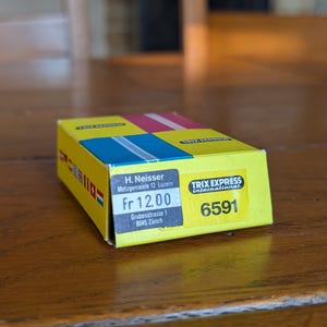 May include: A yellow and multi-coloured box with the text "TRIX EXPRESS international" and the number "6591". The box has blue, pink, and yellow sections. The box also has the text "H. Neisser Metzgerrainle 13 Luzern" and "Grubenstrasse 1 8045 Zürich".