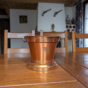 May include: A copper-colored bucket with a flared top and two small handles. The bucket sits on a decorative base with a ring of small, evenly spaced dots. The bucket is on a wooden table.