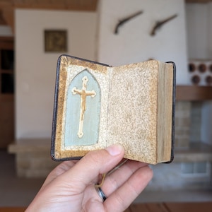 May include: A small, open book with a gold cross on a light blue background. The book has a brown leather cover and aged, patterned pages. The cross is detailed with a gold figure of Jesus. The book is held open, showcasing its contents.